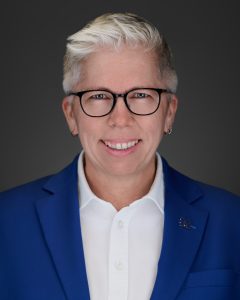 Dr Joël Dickinson in a blue blazer wearing black rimmed glasses and smiling for the camers