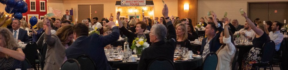 Tourism and Hospitality Management TEAMS fundraising event. People sitting around tables with their hands raised waiting to purchase raffle tickets.