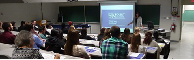 Business students sitting tiered in a classroom facing the front where the instructor is displaying work on a screen.