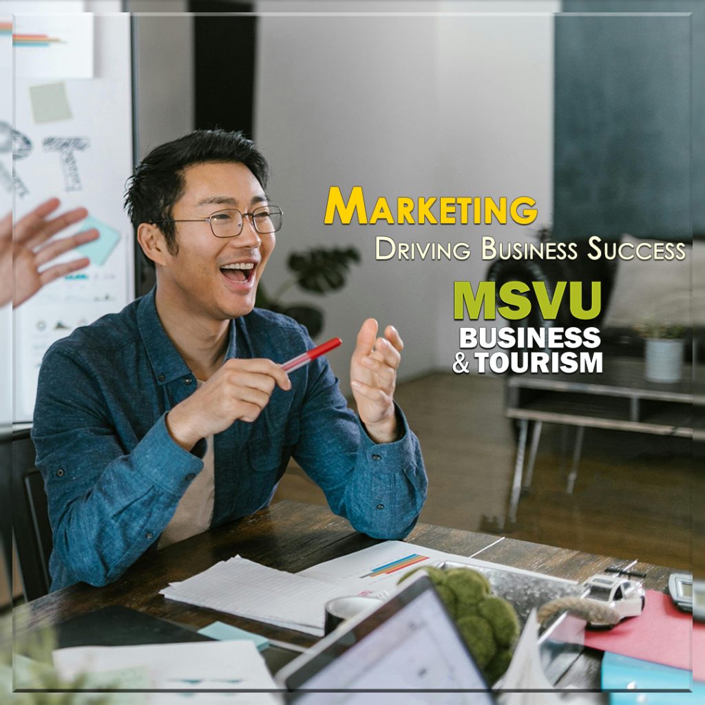 This is a photo of a smiling Asian man who is sitting at a desk with a red pen in his hand. There is a person standing behind and to the left of him and a chart board behind them. They are having an animated happy meeting. Centered in the photo are the words marketing in yellow above driving business success in white and below that is the MSVU business and tourism logo.