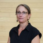 Colleen Blacker, Administrative Assistant to the Department of Business and Tourism. White woman standing in front of wood paneling, in the McCain lobby, smiling, wearing a black short sleeved t-shirt, blond hair pulled back. 