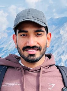 Headshot of Iqbal Singh. Iqbal is a brown man with short dark hair, a short cut mustache and beard. He has a blue ball cap on and appears to be standing in front of a mountain. And he is smiling. 