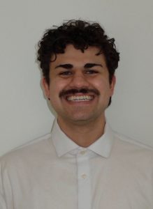 Headshot of Justin who is the recipient of the winter 2026 Pacrim Hospitality Services Inc. Endowed Award for Excellence in Tourism and Hospitality Management. Justin has a huge smile, dark curly short hair, a mustache and a white shirt. He is standing in front of a while background.  