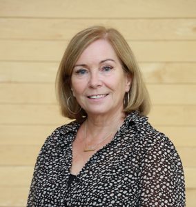 Dr. Sandi Findlay-Thompson, Professor, Management, in the Department of Business and Tourism. White woman standing in front of wood paneling, in the McCain lobby, smiling, wearing a patterned shirt, blond hair. 