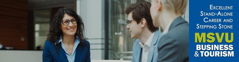 This is a photo of a white woman with dark shoulder length hair and black rimmed glasses. She is smiling and looking at two men in suits. She has on a dark blue blazer and light blue shirt. Beside the image, on the right are the words in white excellent standalone career and steppingstone with the business and tourism logo below. This is on a dark blue background.