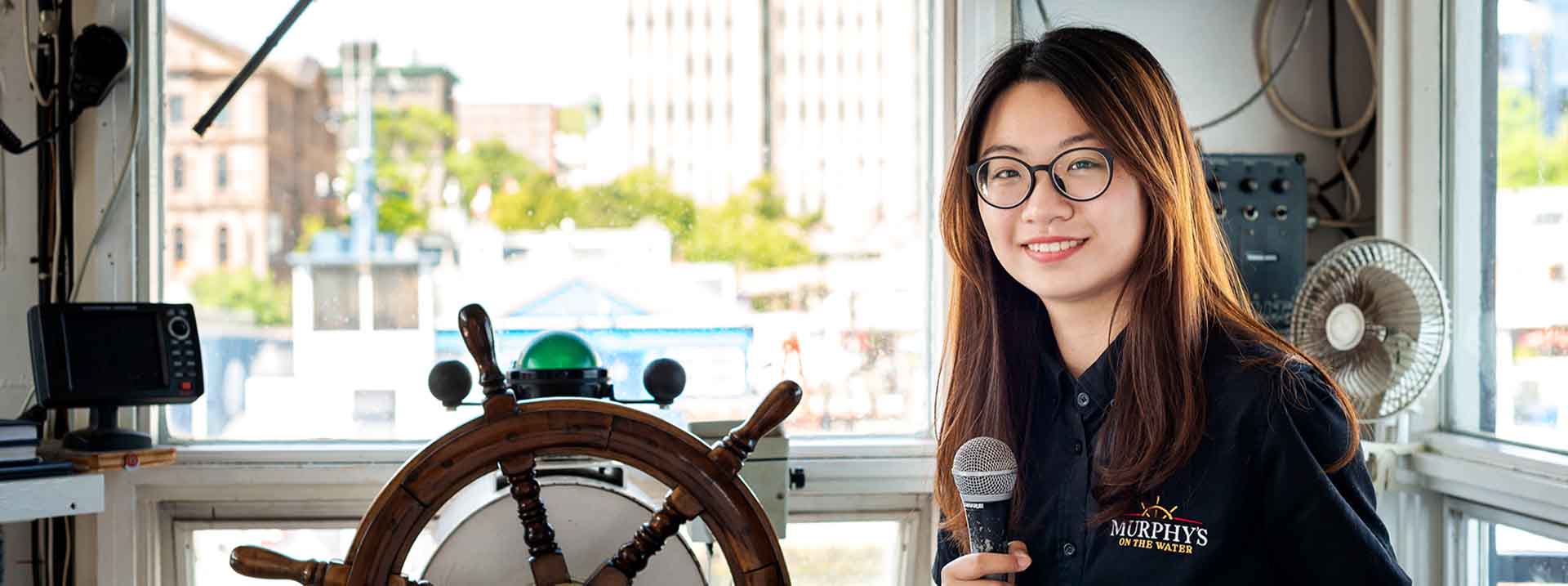 This is a photo of a female tourism and hospitality management student who is on her work term with Murphy's. She is holding a microphone and is in the bow of a tour boat.