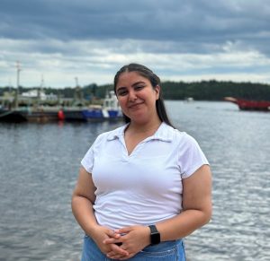 Photo of Jessica Kapoor recipient of the Ann and Jim Duffy award. Jessica is standing in front of the ocean by a fishing boats tied up on a doc. It is a partially cloudy day but Jessica, wearing white t-shirt with collar and jeans, is smiling. 