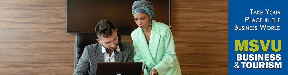 This is a photo of two people in a brown office looking at laptop that is on a desk. A woman with a head scarf wearing a light green blazer is standing showing a man with dark hair and a grey blazer something on a computer. On the right with a dark blue background are the words in white, take your place in the business world which is above the MSVU business and tourism logo.