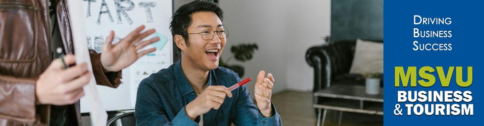 This is a photo of a smiling Asian man who is sitting at a desk with a red pen in his hand. There is a person standing behind and to the left of him and a chart board behind them. They are having an animated happy meeting. to the right, with a dark blue background are the words driving business success in white and below that is the MSVU business and tourism logo.