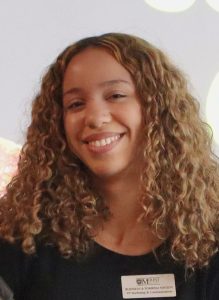 Headshot of Kya Blanch 2026 Ann and Jim Duffy Memorial Award winner in the Business Administration program at MSVU. Kya is a black woman with long brown hair, in a black shirt and is standing in front if a grey background. And she is smiling.