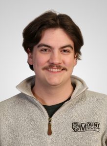 Headshot of Tanner Saunders 2026 Robert (Bob) Bagg Endowed Award winner in the Business Administration program at MSVU. Tanner is a white man with brown hair, in a beige Mount sweater and is standing in front if a grey background. And he is smiling.
