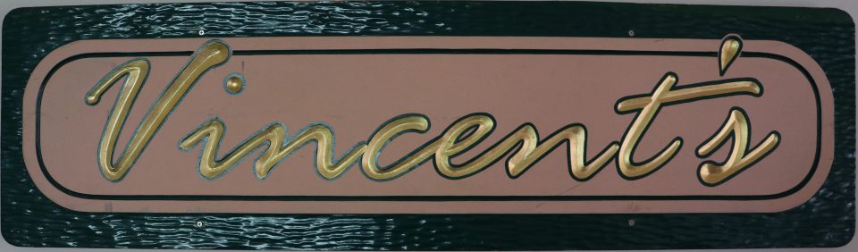 Glossy wood painted black around the edges with the word Vincent's centered in the middle in gold on a beige oval in cursive writing.