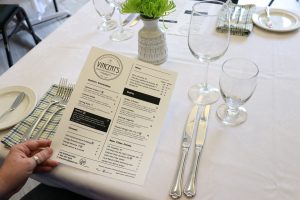 The Mount's Tourism and Hospitality Management student kitchen which is called Vincent's Restaurant. Photo of a hand holding a menu over a fully set table. 