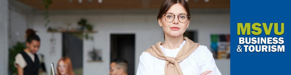 This is a photo of a white woman in the foreground wearing a white shirt with a beige sweater tied over her shoulders. She has brown circular glasses and brown hair tied back. In the background are three people working in an office. On the right is the business and tourism logo on a dark blue background.
