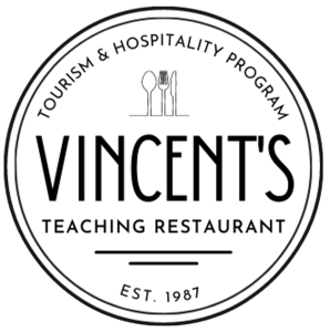This is the MSVU Tourism and Hospitality Vincent's teaching restaurant logo. It is black text on a white background. The logo is circular. At the top it says Tourism & Hospitality Program. Under this is a spoon, a fork and a knife. Under this in big letters is the word Vincent's and under that it says Teaching Restaurant, followed by two lines and under the lines it says est 1987.