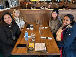Four smiling female business and tourism students with their coats on sitting in a wood booth at Gahan House. They have water glasses and menu's and are getting read to order lunch. 