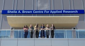 Alt= The front of a building covered in glass windows and has a large balcony. Six people are standing on the balcony facing the camera and leaning with their forearms on the railing of the balcony. Above them, on the front of the building, is a blue sign that says, "Sheila A. Brown Centre for Applied Research." 