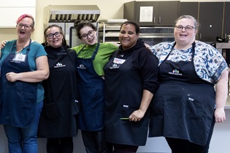 Five students posing for a photo in an industrial kitchen, wearing black aprons and smiling at the camera