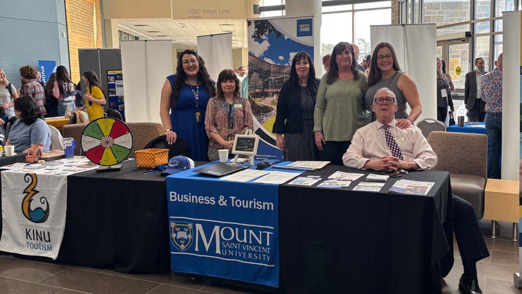Business and tourism faculty and staff at the spring 2026 MSVU open campus day. Five women and one man sitting and standing behind the information table in the McCain atrium.