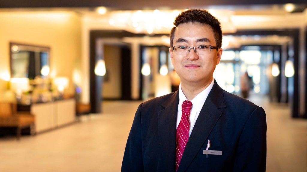 MSVU male tourism and hospitality student on a work placement in a hotel. He has on a dark blue suite with a red tie, and he is smiling whilst standing in front of a hotel lobby.