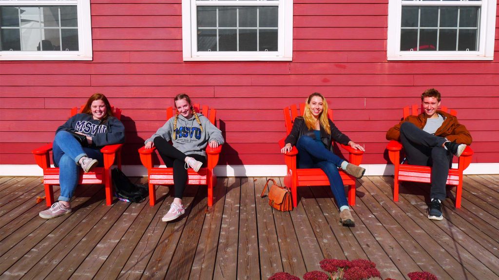 Four MSVU business and tourism students sitting in Adirondack chairs on a patio during one of the sustainable business tours. The chairs are red and the building behind them is a different red with white windows. Three women and one man. Two of the women have MSVU sweatshirts on. They are all smiling. It is a sunny day.