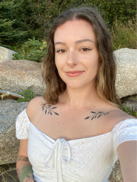 Megan - a white woman with brown hair wearing a white top that shows off vine tattoos on her clavicle.