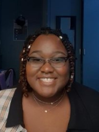 Shermiaya - a Black woman with chin-length braided hair wearing a black and plaid shirt smiling at camera.