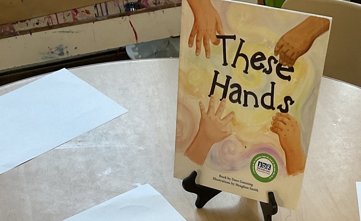 A Book titled "These Hands" on a stand in front of a few sheets of paper for arts and crafts