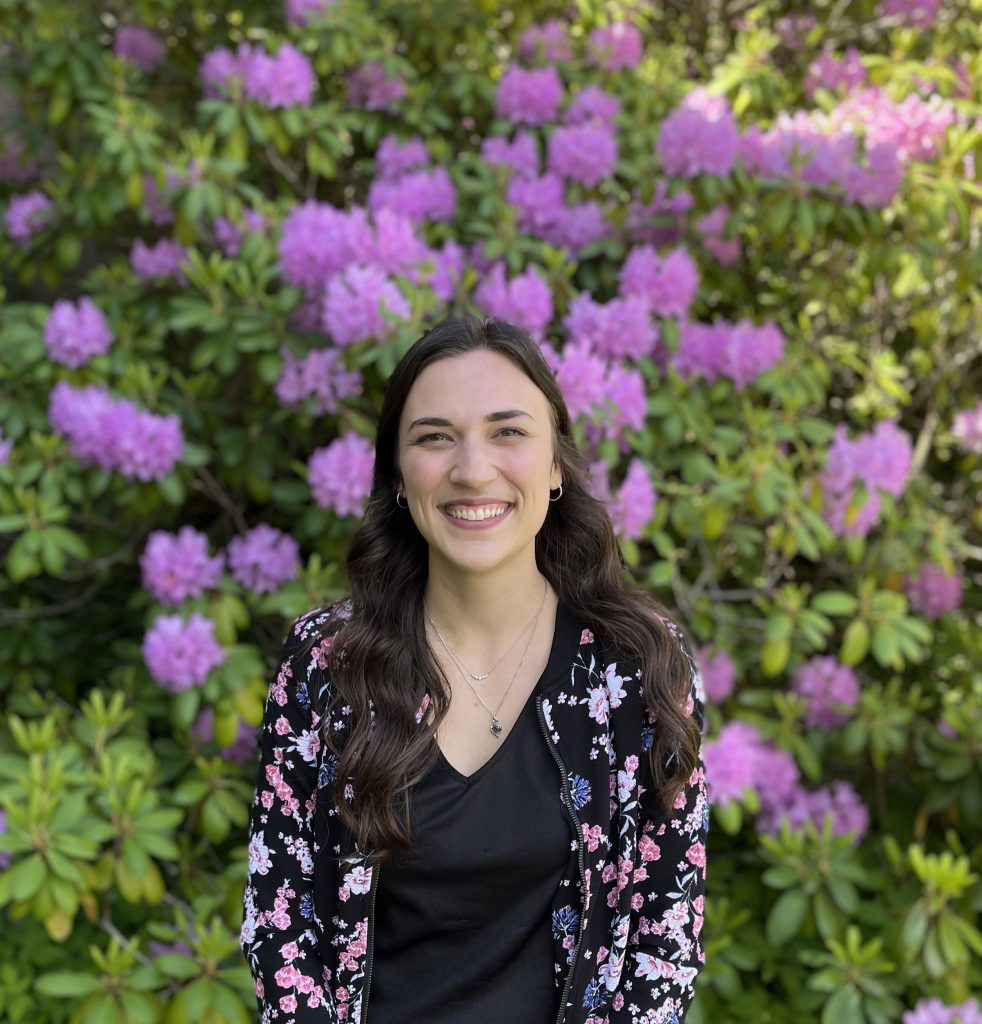 Alt= a person smiling at the camera and the camera frame shows their chest up. They are wearing a black t-shirt and a black jacket with pink, blue, and white flowers all over it. They have mid-length black hair.