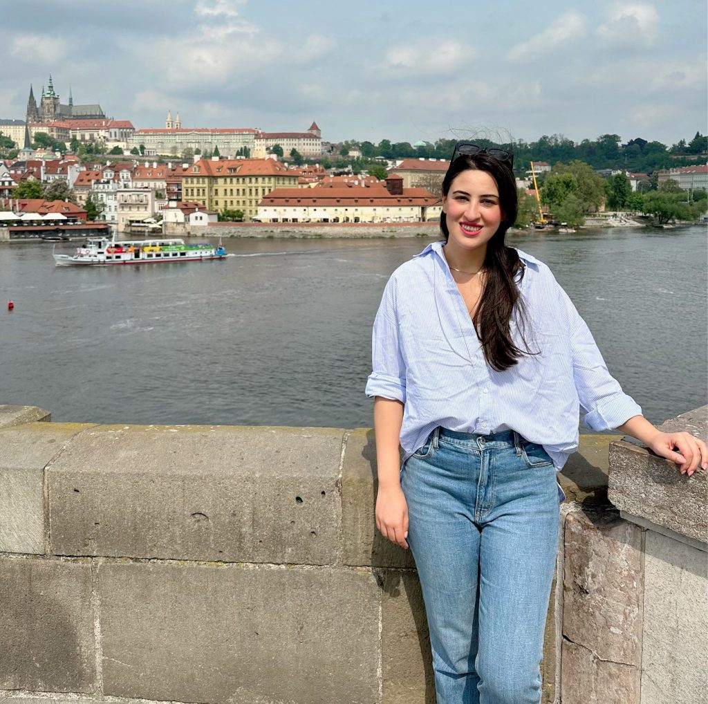 Alt= a person smiling at the camera and the camera frame shows the knees up. They are wearing a light blue button up shirt and light blue jeans. They have black hair that is in a low ponytail. They are leaning against a short wall, in the background you can see a canal with a boat.