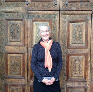 Alt= a person smiling at the camera. They are wearing a blue pullover, a coral scarf, and black pants. They have their hands clasped in front of them. Their hair is in a ponytail and their hair is silver.