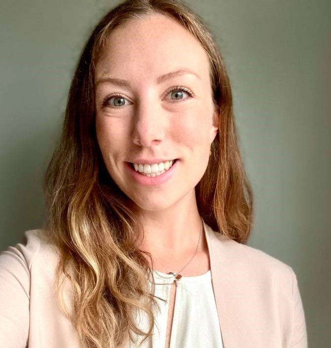 Alt= a selfie of a person smiling and the frame is shoulders up. They are wearing a white blouse and a pale pink blazer. Their hair is mid-length and brunette.