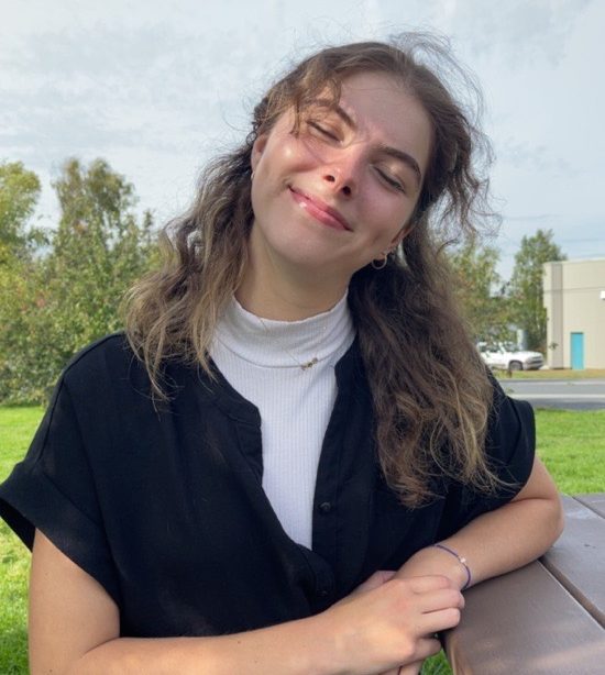 A woman with long brown hair is sitting at a picnic table. She is wearing a black button up short sleeve shirt over a white shirt and smiling at the camera