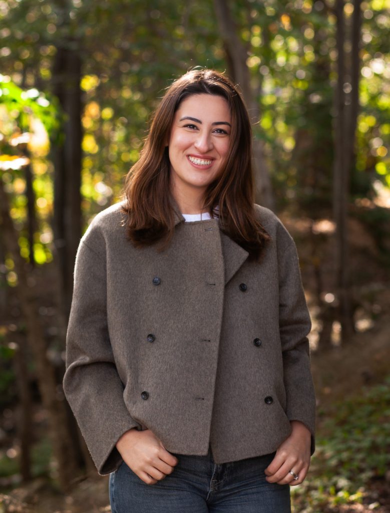 A woman with short brown hair smiles into the camera. She is wearing a waist-length grey coat.