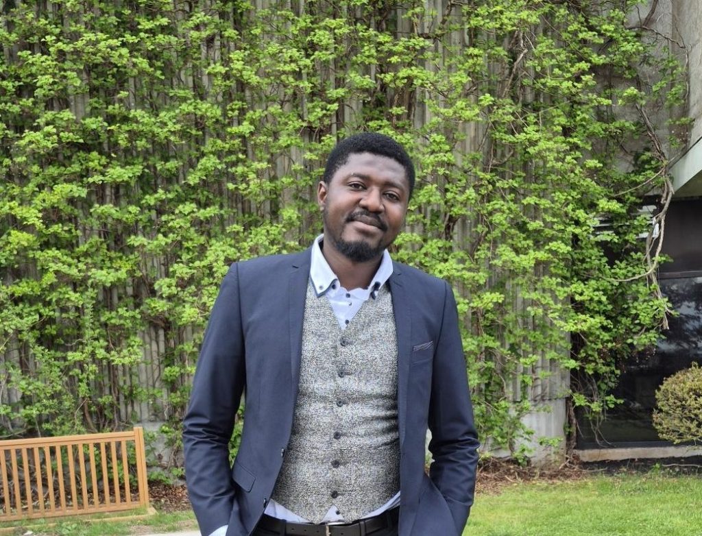 A black male adult in a suit with his hands in his pocket, smiling into the camera