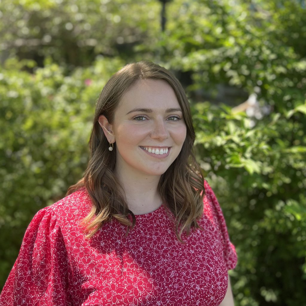 Alt= a person smiling at the camera and the camera frame shows shoulders up. They are wearing a red t-shirt and their hair is shoulder-length and brunette.