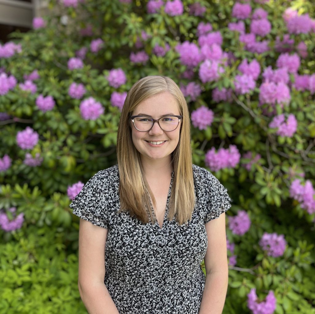 Alt= a person smiling at the camera and the camera frame is chest up. They are wearing a black t-shirt with many small white spots on it. Their hair is mid-length and blonde. They wear glasses.