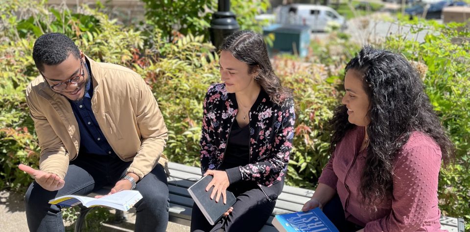 Alt= three people sitting on a bench with a green bush behind them. The person on the left is reading a book. The other two people beside the reader are holding closed books and looking at the book in the hand of the reader.