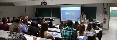 Students sitting tiered in a classroom facing the front where the instructor is displaying work on a screen.