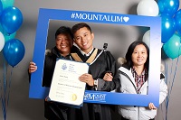 A Business and Tourism student after graduation holding their parchment with their family in the reception area with a holdup blue frame that says Mount Alumni on it. They are surrounded with blue and white balloons. 