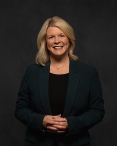 Angela Lewis, Chief Commercial Officer, Stewart McKelvey. Business and Tourism Fireside Chat series 2025. Angela is a white woman with blond shoulder length hair, she is wearing a black shirt under a green grey blazer, she is smiling with her hands held together in front of her. The back ground is a deep grey. 