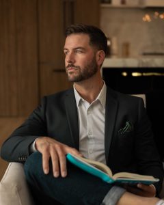 Dave Reynolds, CEO, The Rumin8 Group Inc. Business and Tourism Fireside Chat series 2025. Dave is a white man with brown short hair and a beard. He is wearing a white shirt with a color under a grey blazer. Dave is sitting, holding a book on his lap and looking to the left. 