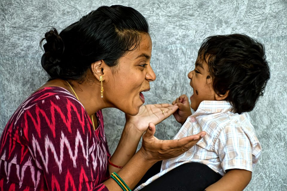 Alt= an adult and toddler facing each other. The adult is making an o-shape with their mouth and holding their hands up. The child is smiling.