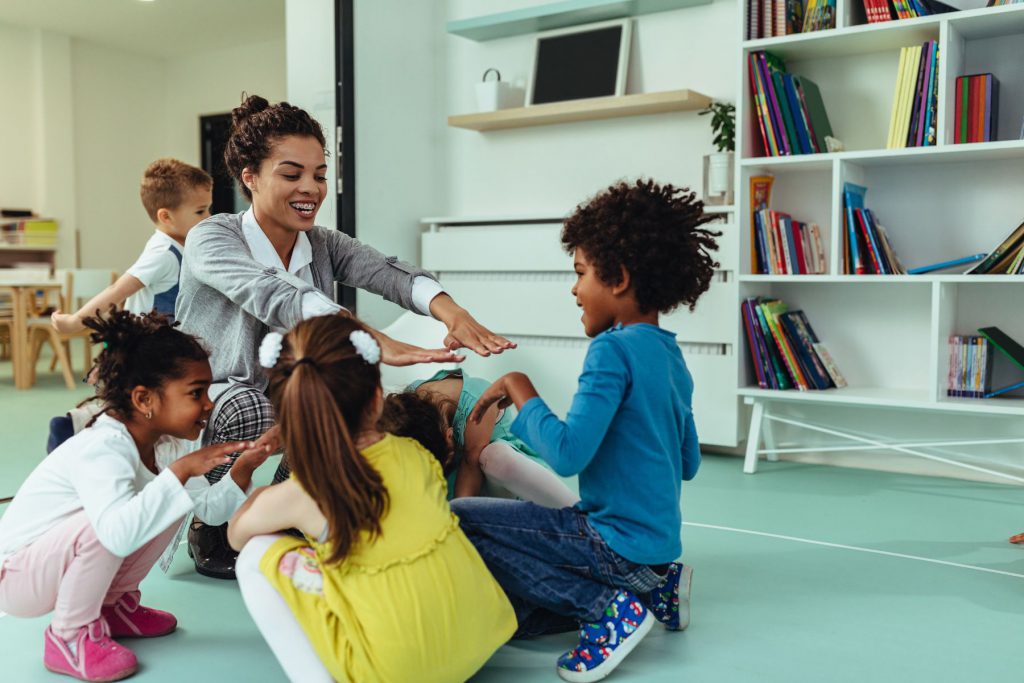 Alt= an adult sitting on the floor with three children sitting around the adult. The adult is reaching her hands out to one of them