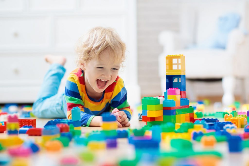 Alt= a child laying on their stomach smiling surrounded by various coloured blocks