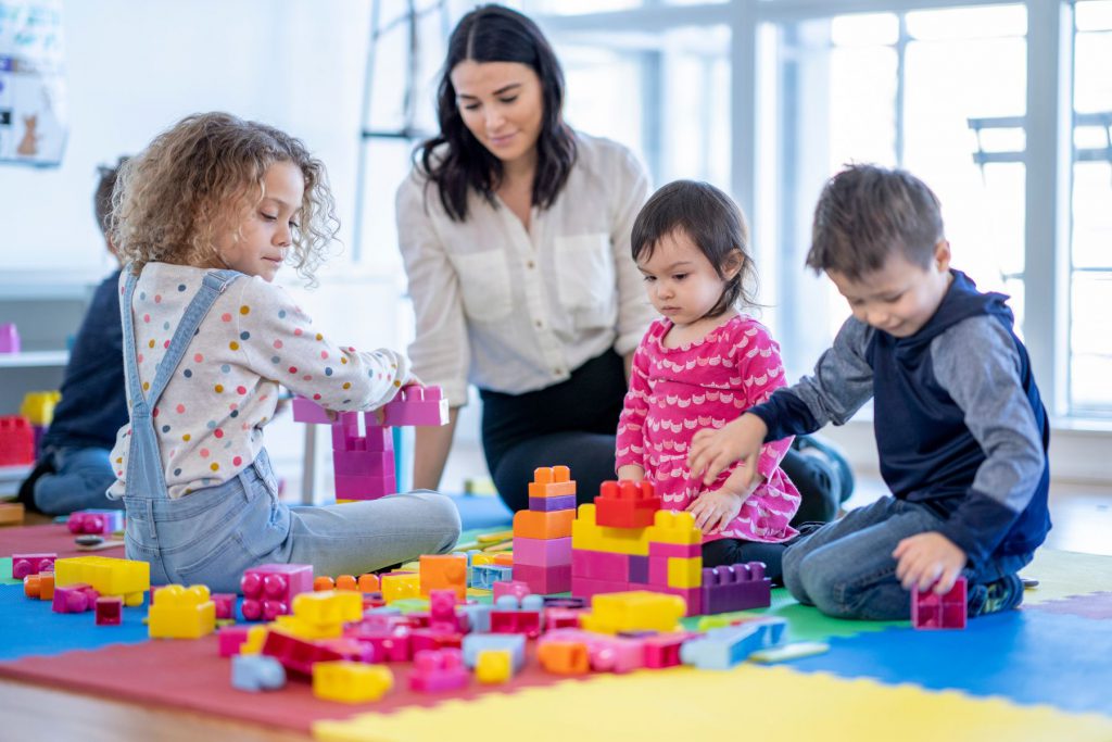 Alt= an adult and three children sitting on a playmat that is covered in various coloured blocks. The adult and one of the children are looking down at the blocks in front of them. Another child is looking at the block they are holding in their hand. The third child is looking at their arm