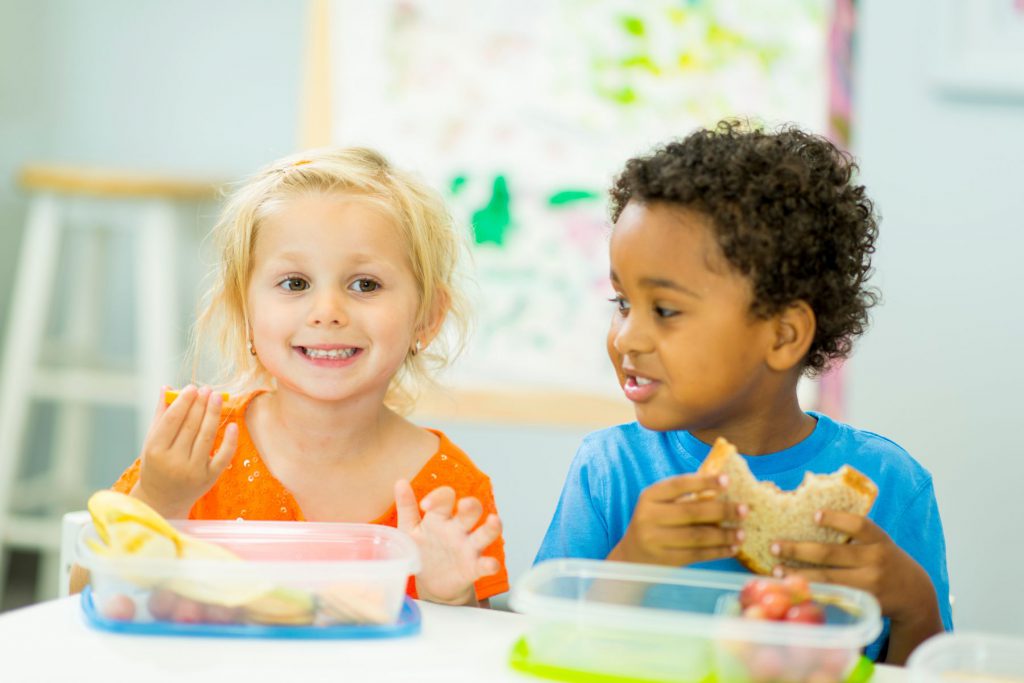 Alt= two children eating food out of plastic container. One child is smiling at the camera with an orange food in their hand. The other child is smiling at the first child mentioned and this child has a sandwich with a big bite taken out of it in their hand