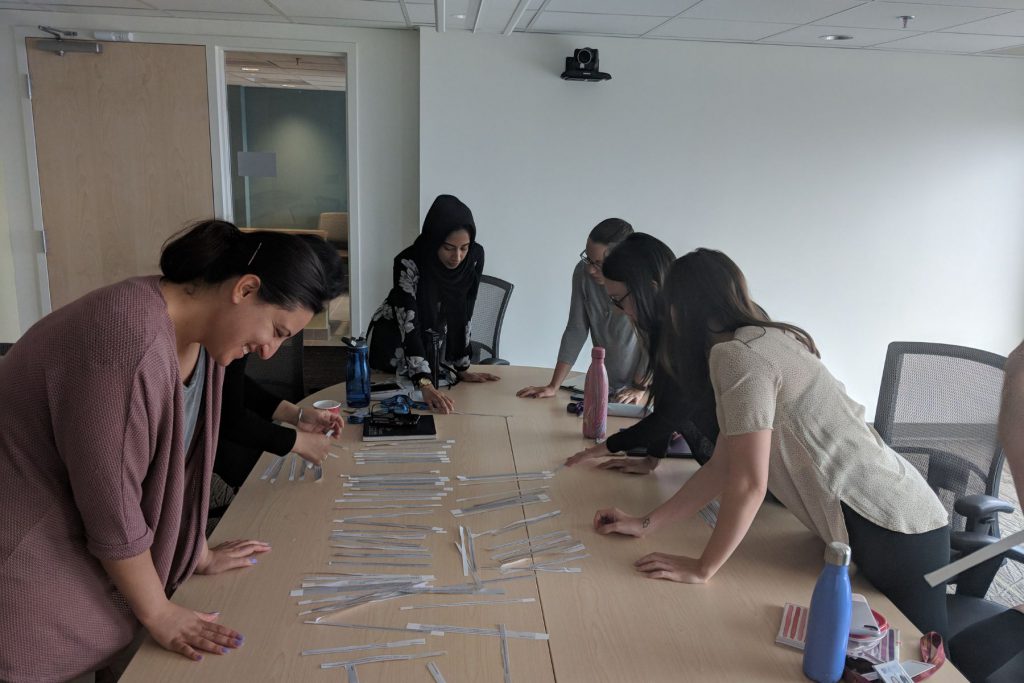 Alt= five people leaned over a long rectangular table all looking down at strips of paper spread out over the table.