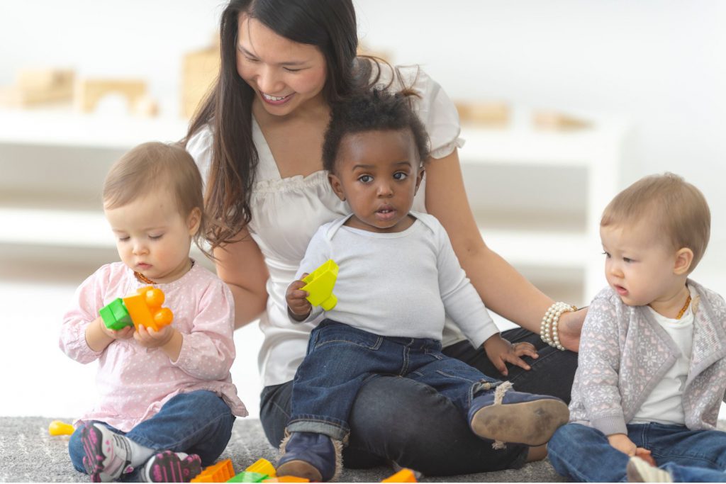 Alt= an adult sitting on the ground with three toddlers. One toddler is on the adult's lap holding a block. One is sitting to the left of the adult with two blocks and one is sitting to the right of the adults looking at the toddler on the left. The adult is smiling down at the toddler on the left.