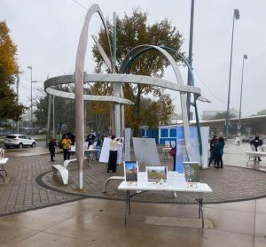 Alt= a shoelace sculpture at the Emera Oval in Halifax Nova Scotia that is shaped like a dome. Under and around the sculpture are tables with three images sitting up on them. There are a few people standing amongst the tables